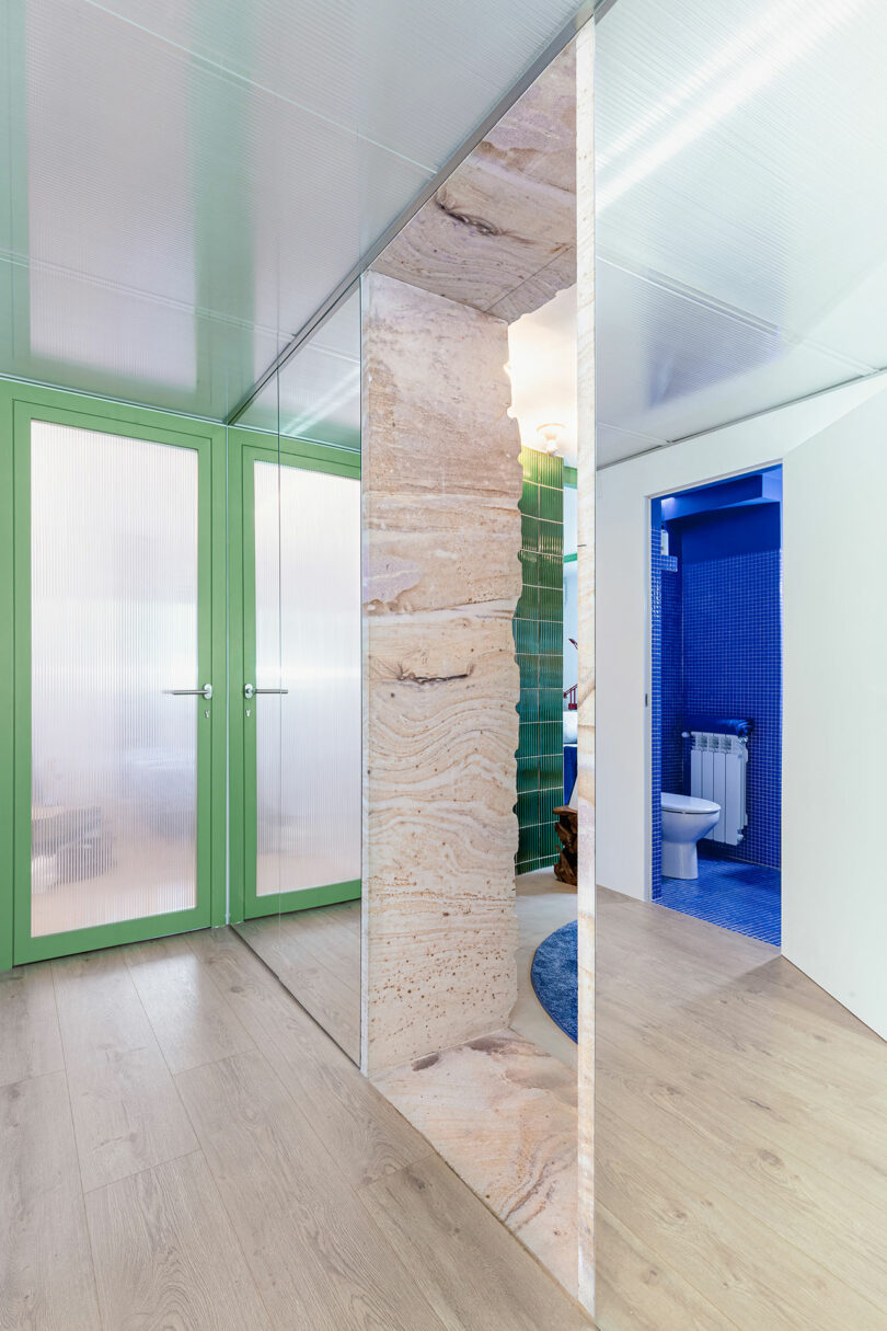 A modern hallway with light wood flooring, green-framed glass doors, and a partition with a natural stone texture. A glimpse of a blue-tiled bathroom is visible through an open door.