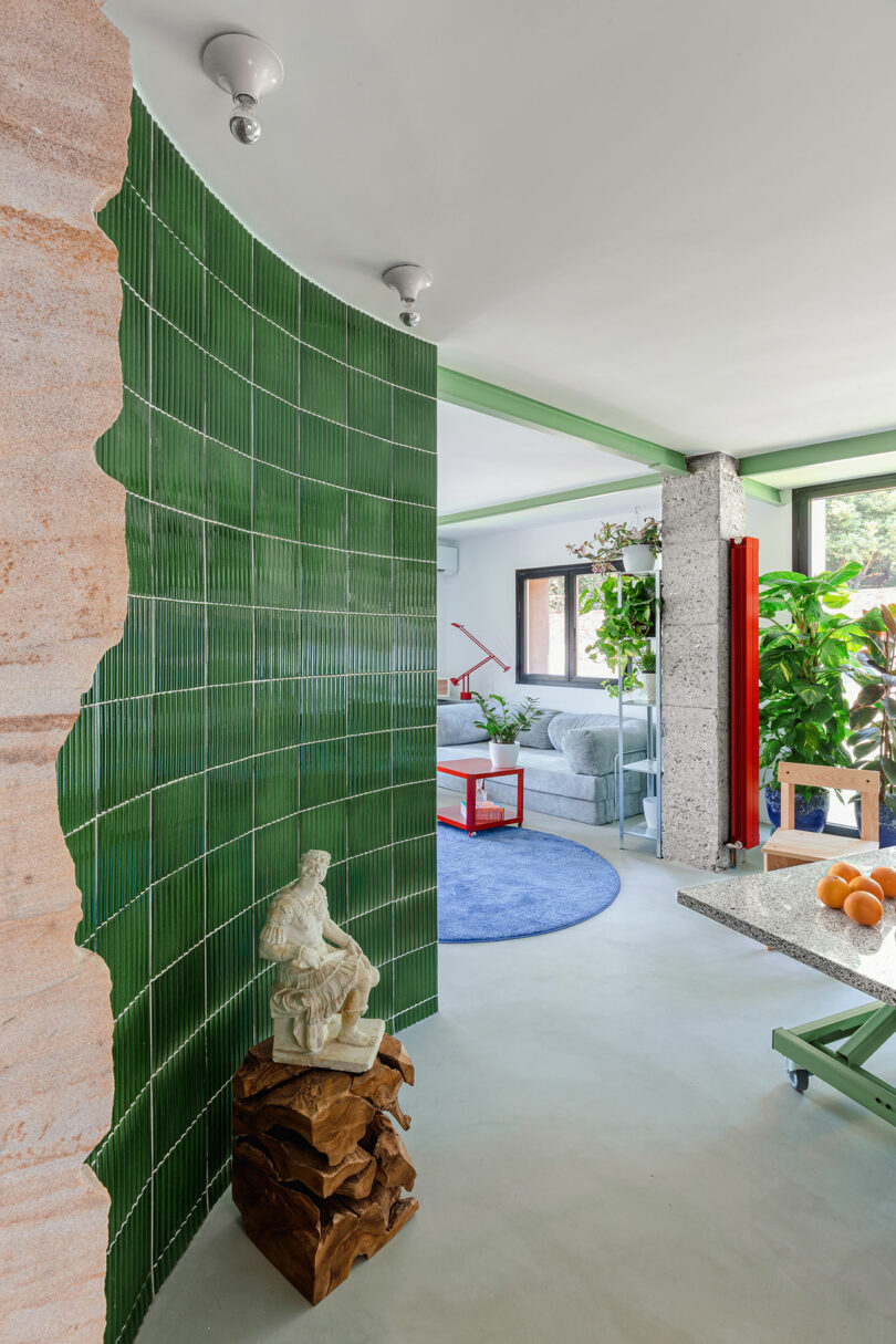 Interior scene with a green tiled wall, a statue on a wooden base, a round blue rug, and modern furniture. Large plants are near windows, and a bowl of oranges is on the table.