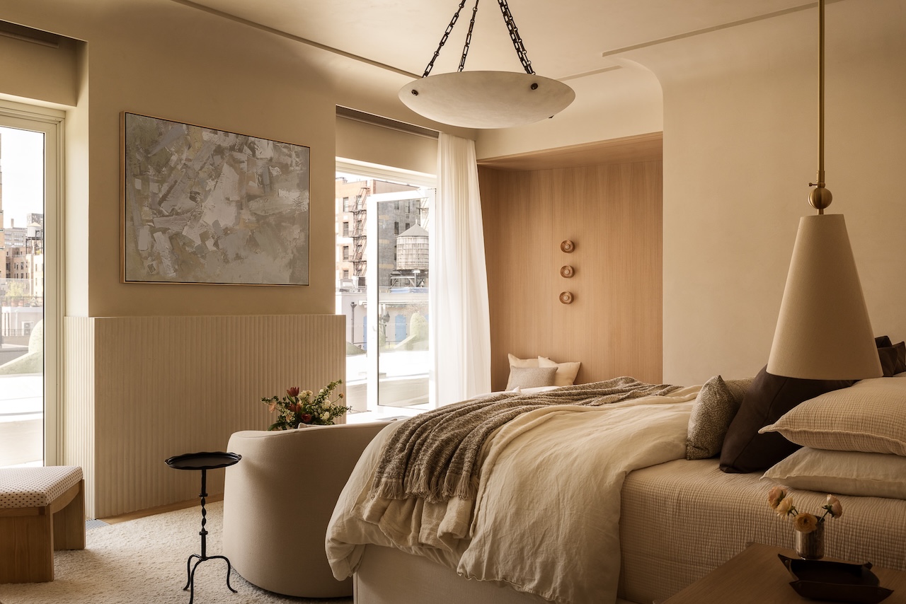 A modern bedroom with beige walls, a large bed with layered bedding, abstract wall art, and natural light streaming in from windows. Minimalist decor and soft, neutral tones throughout.