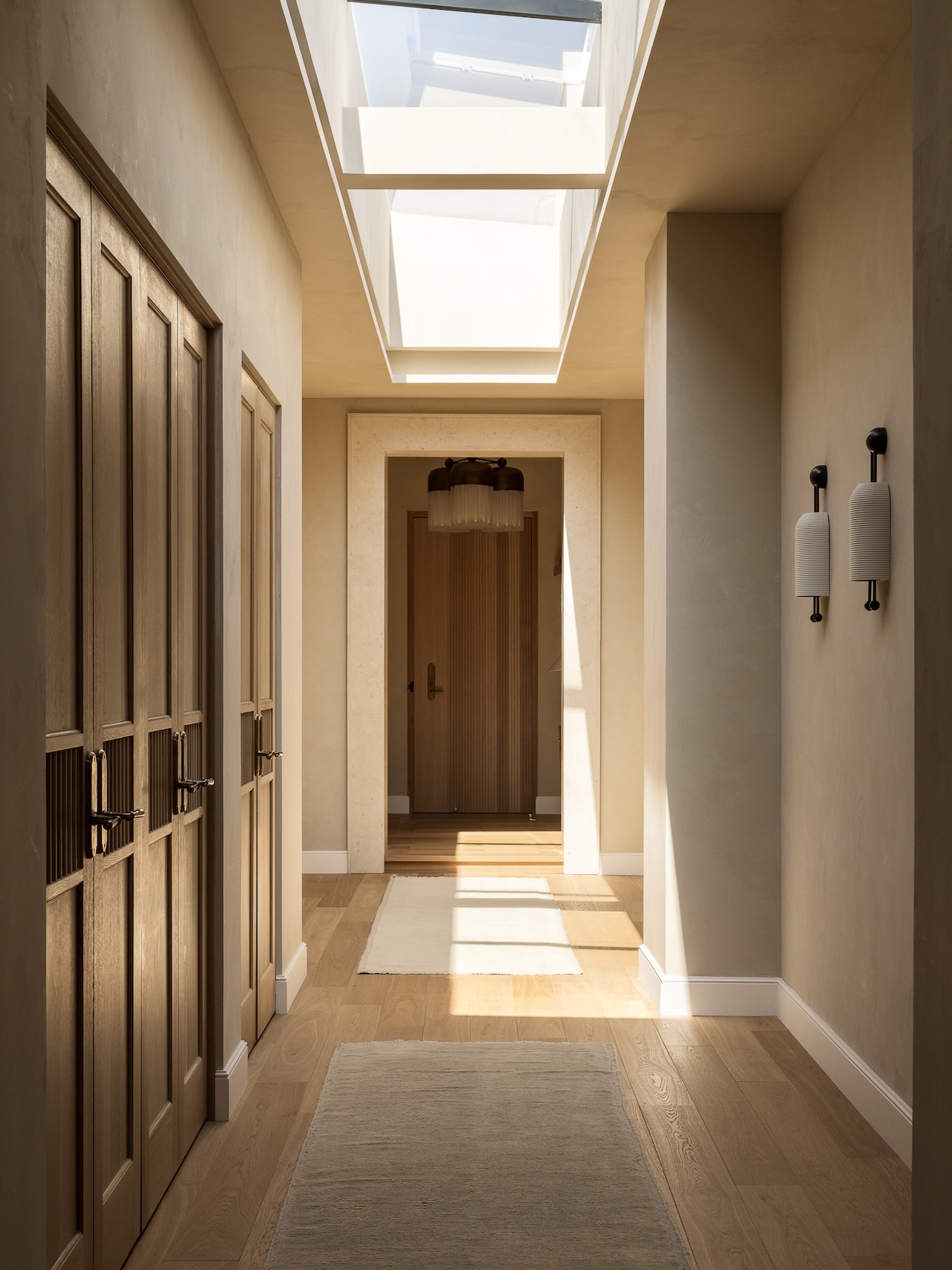 A bright hallway with wood floors, beige walls, skylight, double doors on the left, wall sconces on the right, and a wooden door at the end.