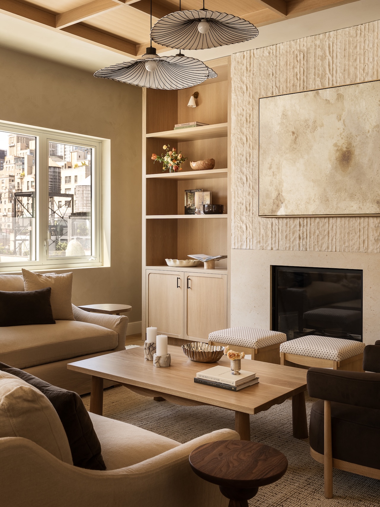Modern living room with neutral tones, a large artwork above a fireplace, built-in shelves, light wood furniture, a window with a city view, and two pendant lights overhead.