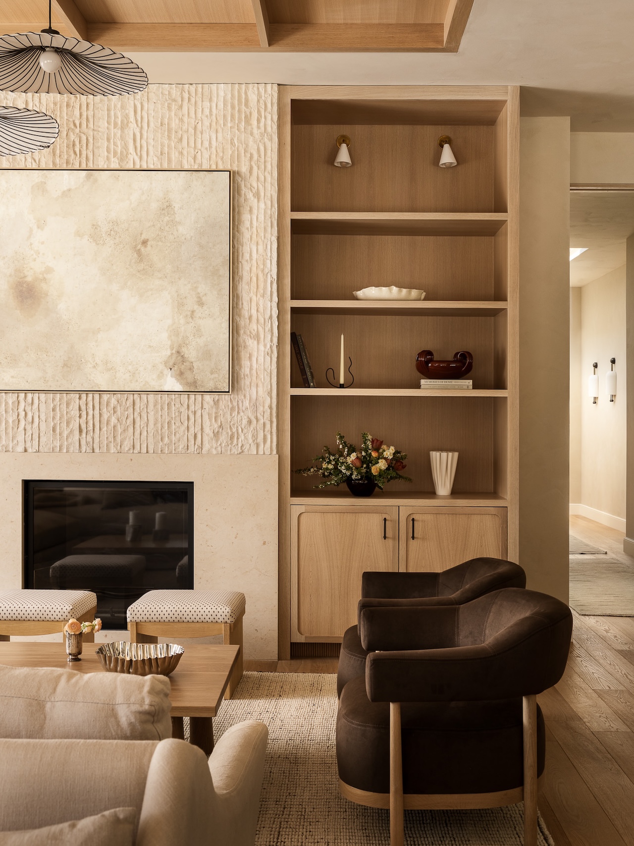 A modern living room features a fireplace, built-in shelves with decor, a dark armchair, a beige sofa, and a light wood coffee table on a textured rug.
