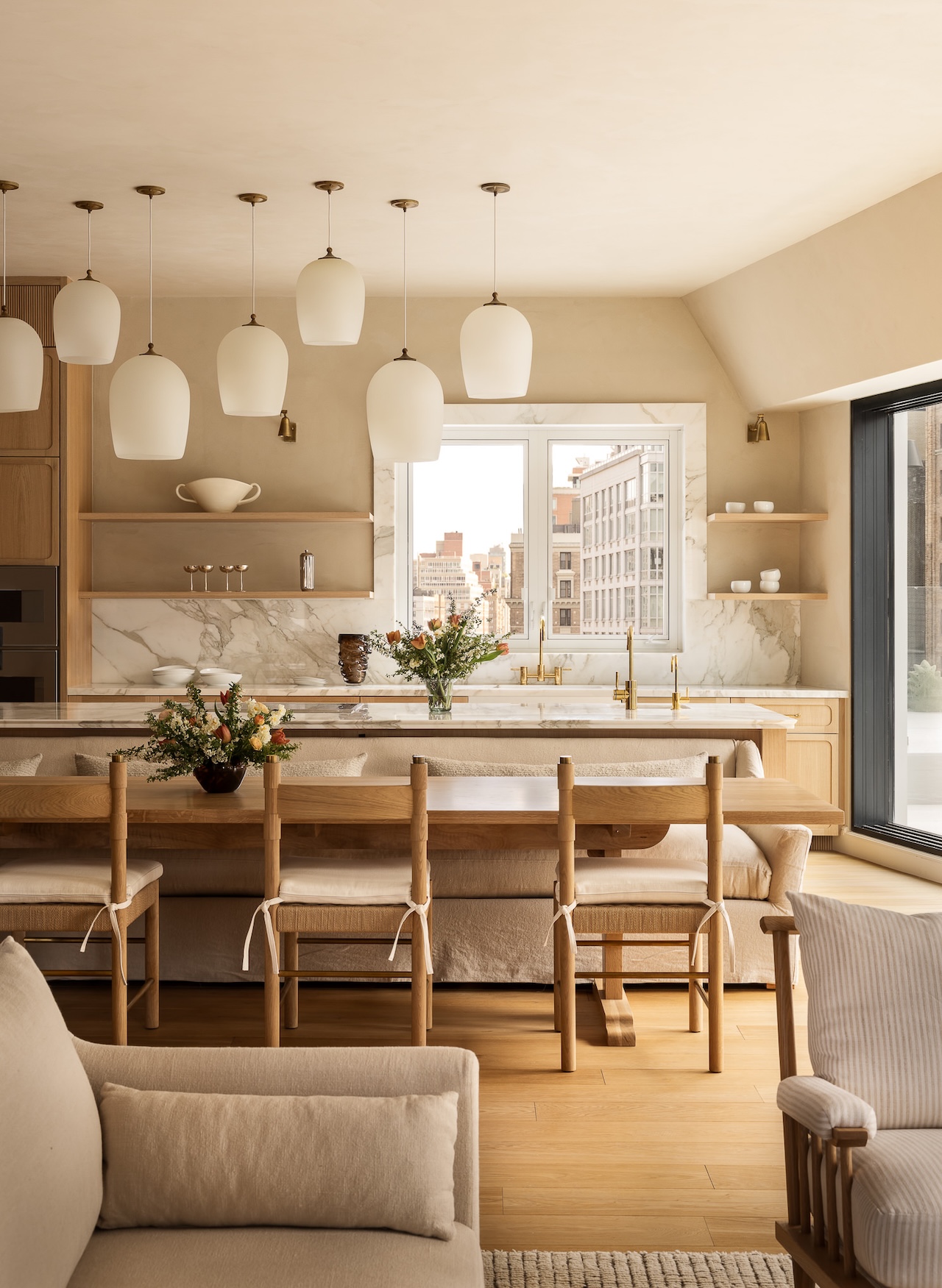 Modern kitchen with wood finishes, marble countertops, hanging pendant lights, open shelves, and large windows showing a cityscape in the background. Dining table with chairs in the foreground.