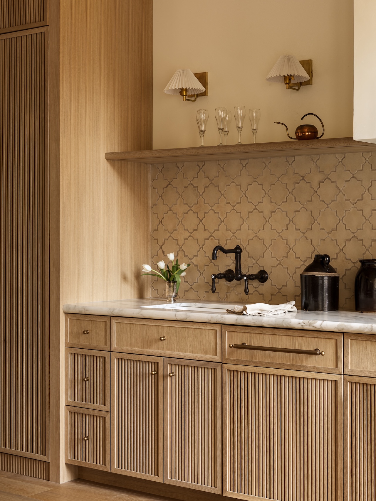 Light wood kitchen cabinets, marble countertop, patterned tile backsplash, black faucet, two black jars, a vase with flowers, glasses on a shelf, and wall sconces above.