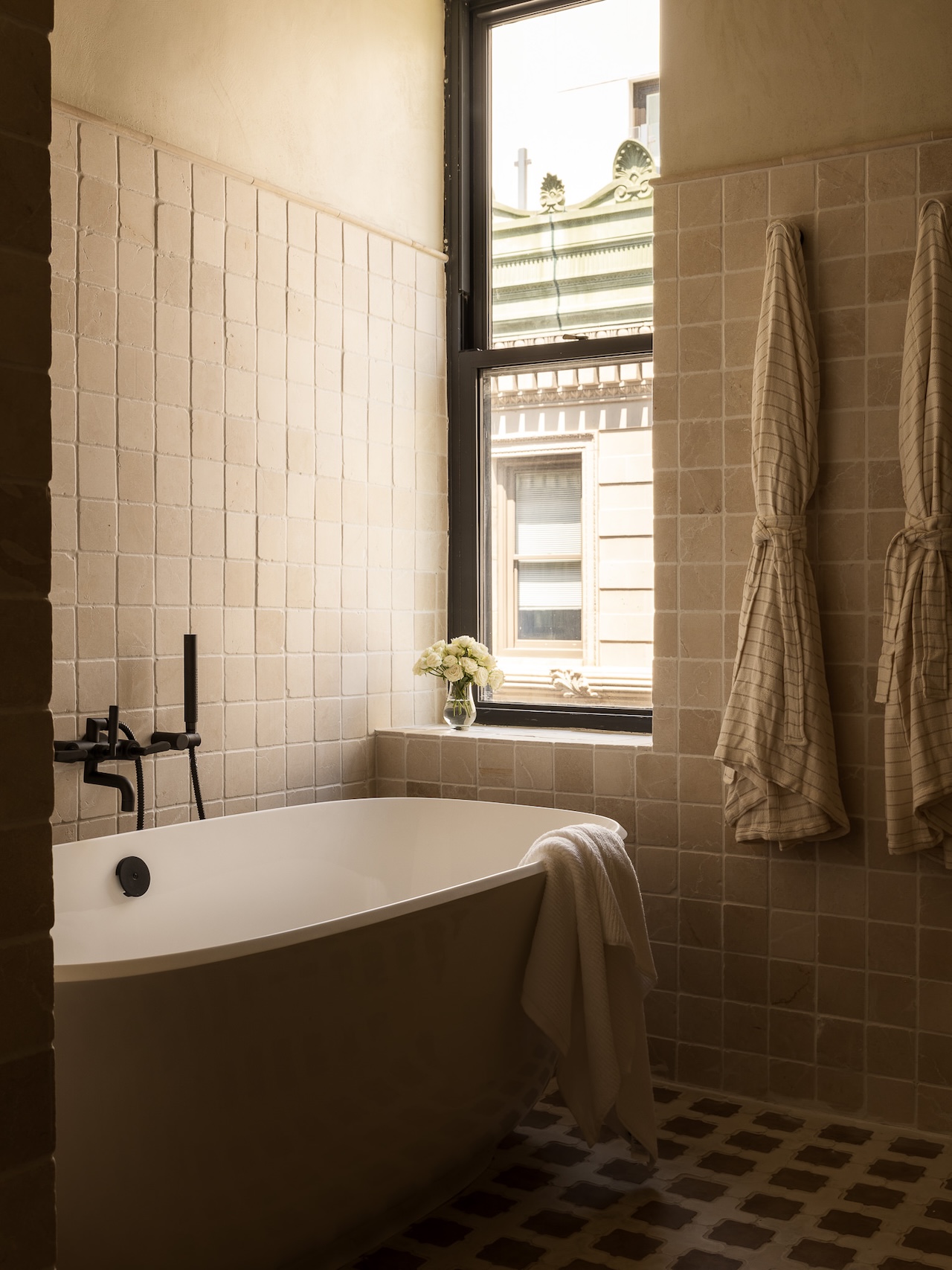 Modern bathroom with a freestanding tub, black fixtures, two towels hanging on the wall, and a window with a city view. A small vase with flowers sits on the windowsill.