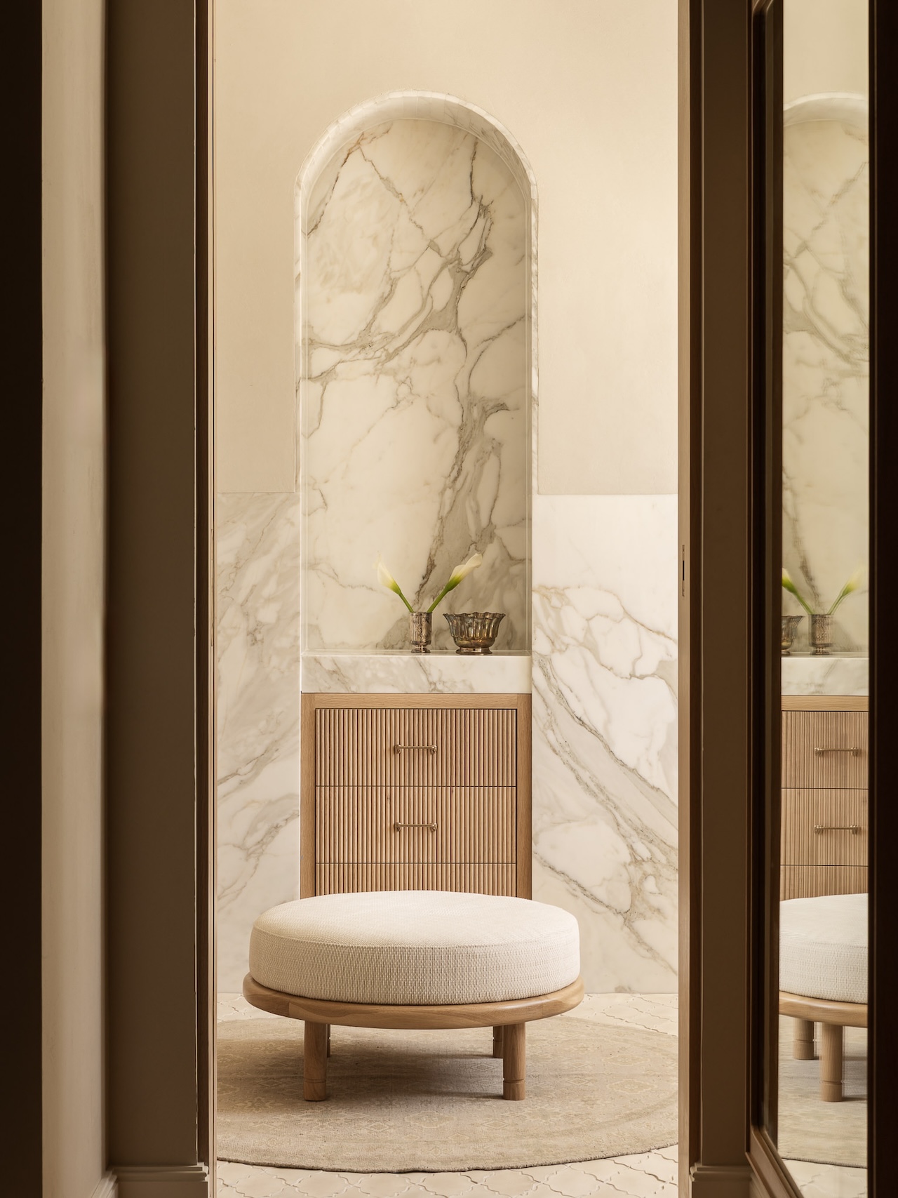 A round upholstered bench sits in front of a wood cabinet with marble accents, two vases with calla lilies, and a decorative bowl in a softly lit, neutral-toned room.