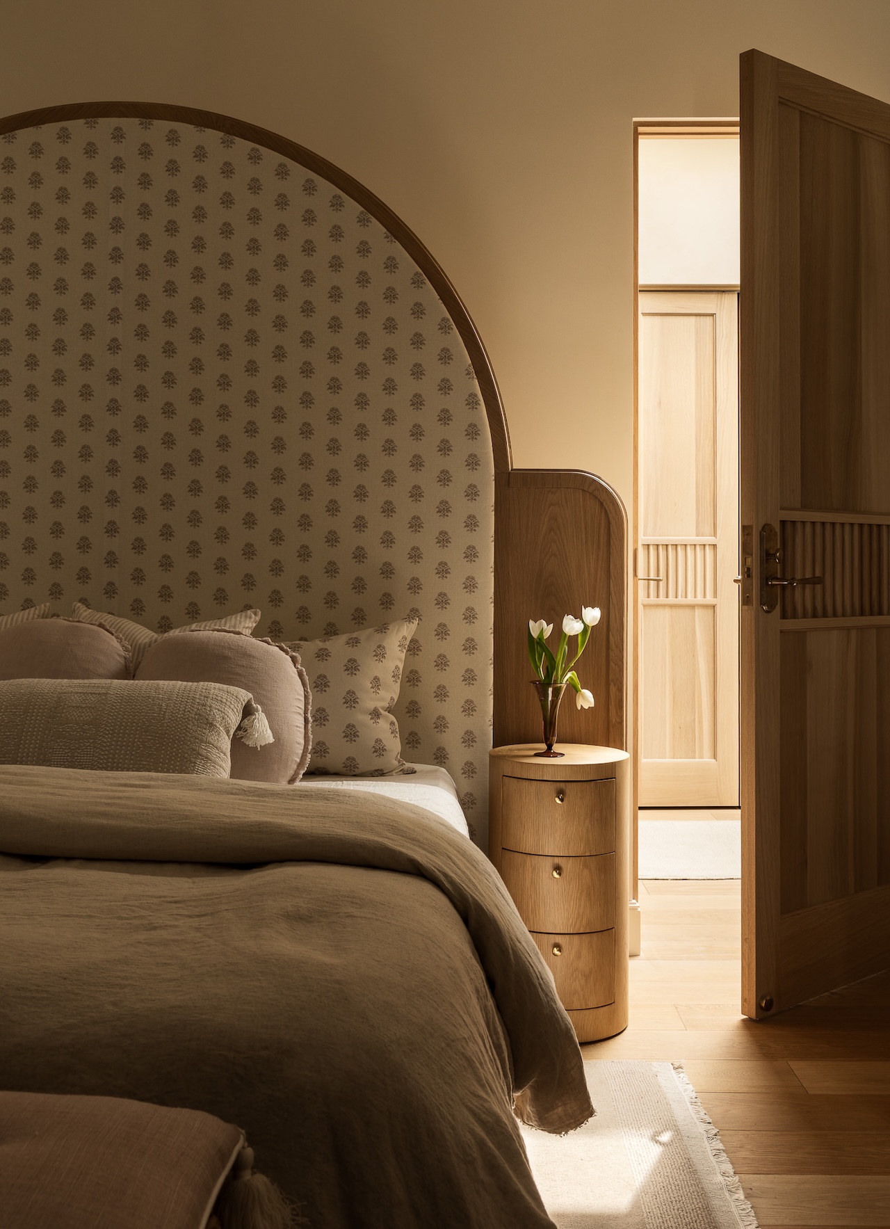 A softly lit bedroom features a curved headboard, patterned wallpaper, neutral bedding, a round wooden nightstand with a vase of tulips, and an open wooden door leading to a light hallway.