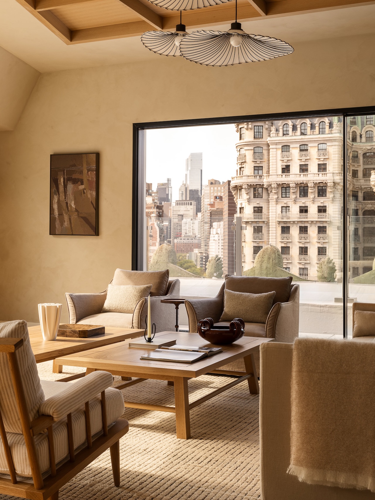 A modern living room with neutral-toned furniture and decor, large window with cityscape view, wooden coffee table, and beige carpeting.