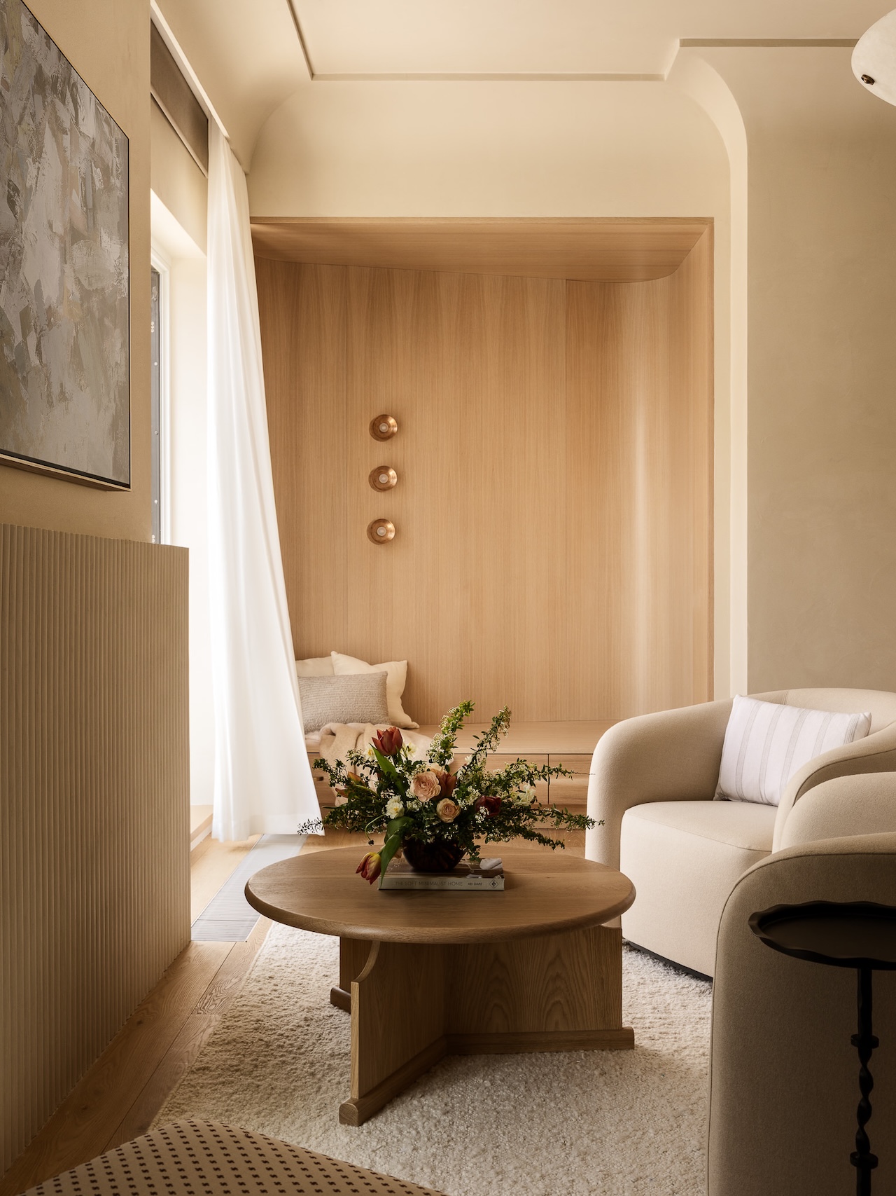 Minimalist living room with neutral tones, featuring a round wooden coffee table, modern armchairs, floral arrangement, large window with sheer curtains, and wall with decorative plates.