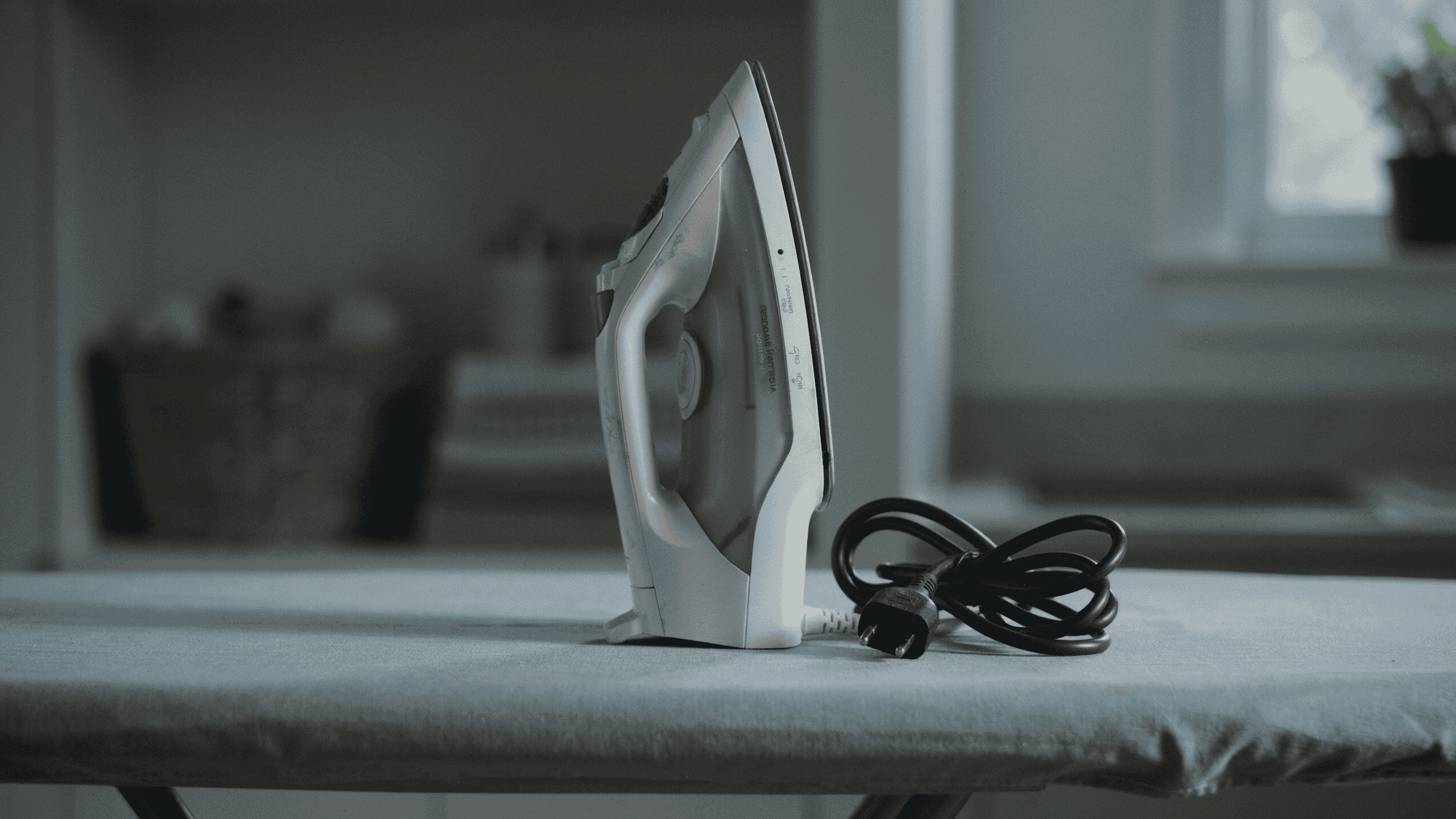 A white electric clothes iron standing upright on a blue ironing board, with its black power cord loosely coiled beside it in a softly lit home interior.